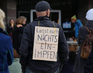Ein glatzköpfiger Herr mit dunkelgrauer Schildmütze trägt ein Schild auf dem Rücken, auf dem steht: "Lasst euch NICHTS EINIMPFEN"