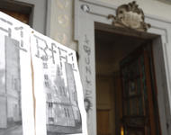 Ein Schild mit Veranstaltungsplakaten der Biennale für Freiburg 1 vor dem Eingang ins historische Klohäuschen.